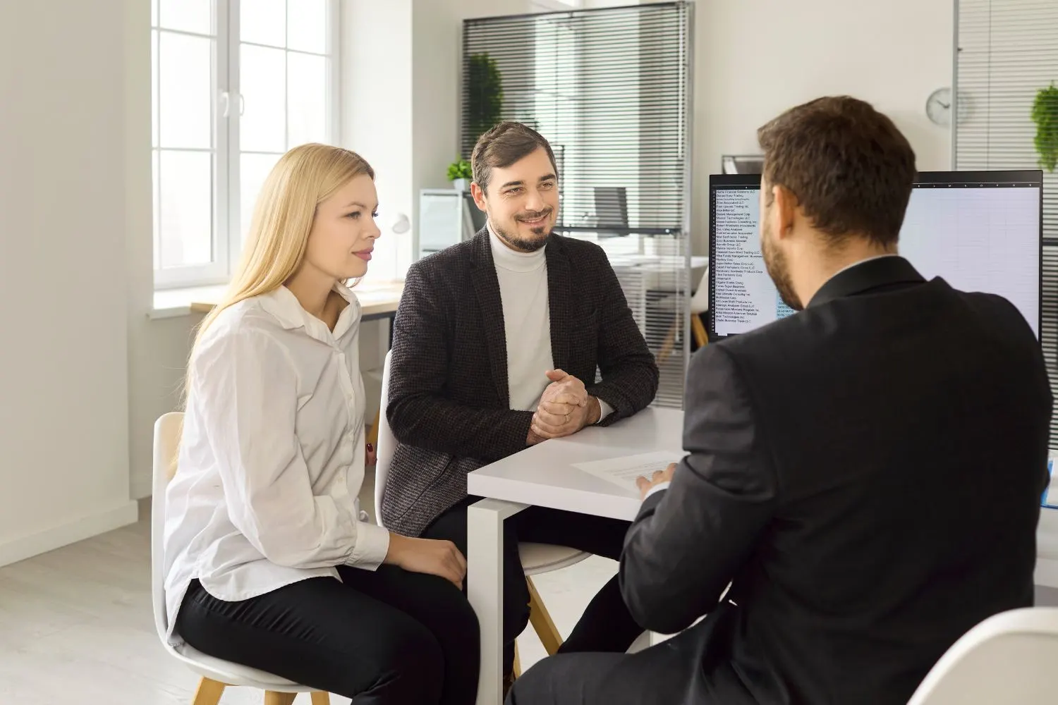 análise de crédito imobiliário em reunião de negócios: corretor e clientes conversam em escritório com mesa de escritório e computador ao fundo, demonstrando avaliação financeira