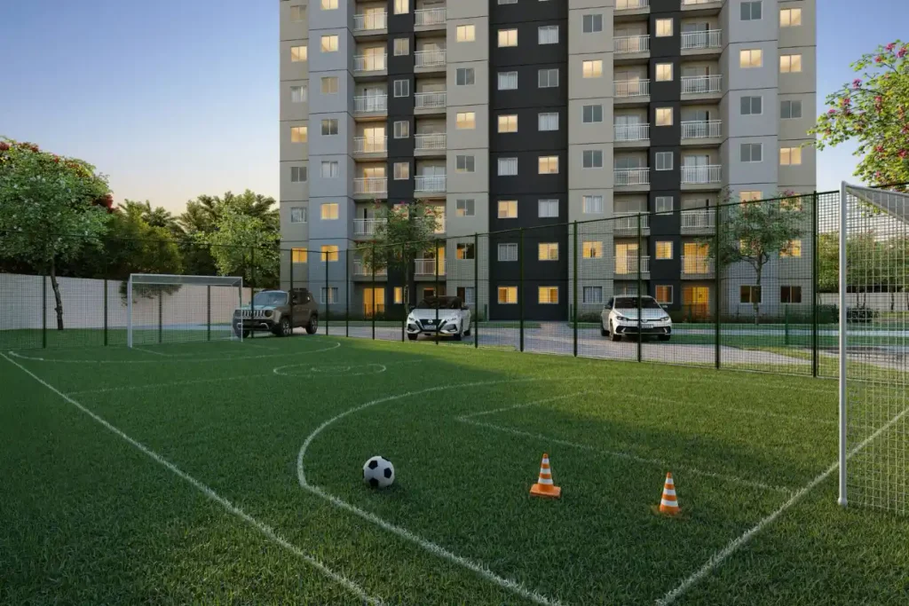 Como comprar um apartamento pelo Minha Casa Minha Vida em um condomínio com apartamentos e área externa com campo de futebol e cerca ao redor, em vista do prédio ao entardecer.